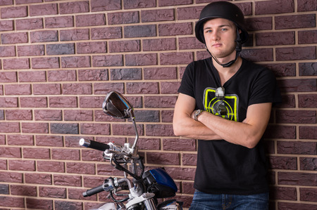 Young Man with Arms Crossed Wearing Helmet Leaning Against Brick Wall and Standing Next to...