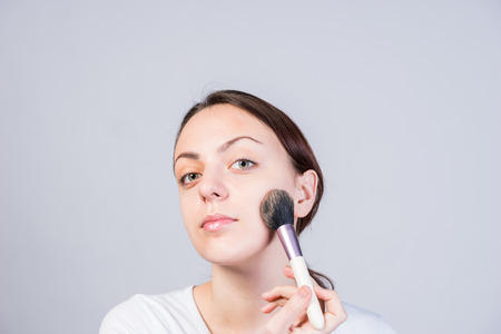 Close up Pretty Young Woman Applying Makeup on her Cheek Using a Brush While Looking at the Camera.