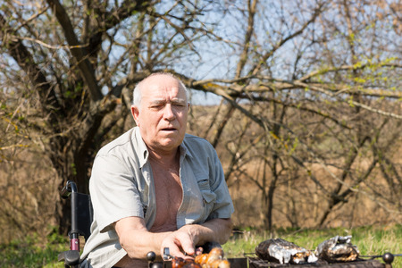 Serious Old Man Sitting on his Wheelchair Roasting Meat Sausage for his Lunch at the Camp Area on a Very Sunny Dayの写真素材
