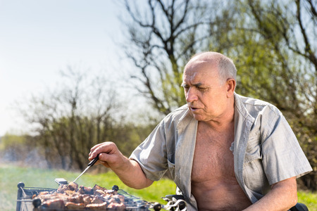 Close up Serious Old Man with Open Shirt Grilling at the Camp Area While Sitting on his Wheelchair on a Very Sunny Morning.の写真素材