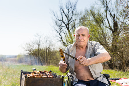 Elderly Man with Unbuttoned Shirt and Sitting on his Wheelchair Holding Grilled Meat on Stick and Knife and Looking at the Camera While Grilling at the Park.の写真素材