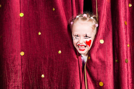 Pretty excited little blond girl on stage waiting to make her entrance on stage during a performance peeking out between the curtains and laughingの写真素材