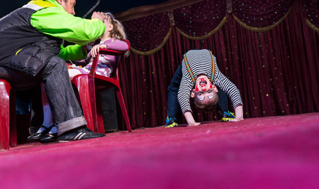 Playful boy with clown make up sitting in bridge position on the stage while his colleague is getting ready with cosmetics for the performanceの写真素材