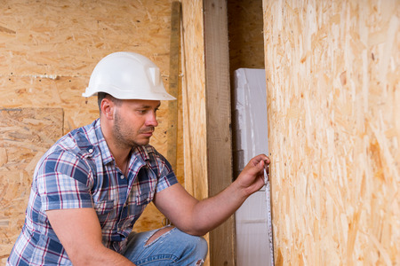 Male Construction Worker Builder Wearing White Hard Hat Measuring Door Frame with Tape Measure Inside Unfinished Home with Exposed Particle Plywood Boardsの写真素材