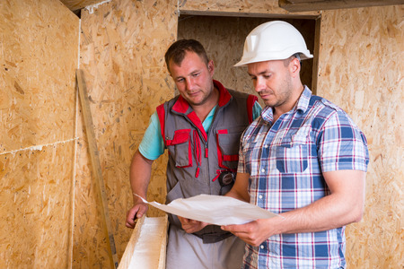 Foreman Wearing White Hard Hat and Worker Looking at Building Plans Together Inside Unfinished Frame...