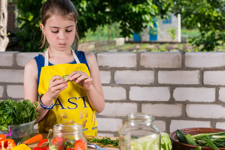 Young girl wearing a yellow apron standing at a table preparing a large assortment of farm fresh vegetables for bottling in glass jarsの写真素材