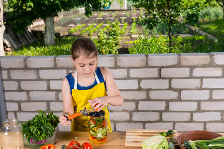 Young girl helping to bottle fresh vegetables standing at a wooden outdoor table carefully placing the veggies in the glass jarsの写真素材
