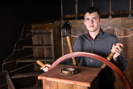 Young Ambitious Business Man Wearing Striped Shirt and Standing at Helm of Ship, Steering Boat with...