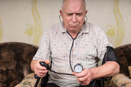 Disabled frowning senior man monitoring his blood pressure through auscultation with a stethoscope...