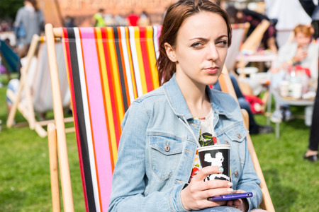 Casual Portrait of Young Brunette Woman Wearing Denim Jean Jacket and Holding Cell Phone and Take Out Coffee Cup While Sitting in Striped Beach Chair in Field at Picnic or Outdoor Festivalの写真素材