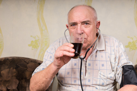 Close up Sick Adult Bald Man with BP Apparatus, Drinking a Glass of Herbal Juice Drink and Looking Straight at the Camera.の写真素材
