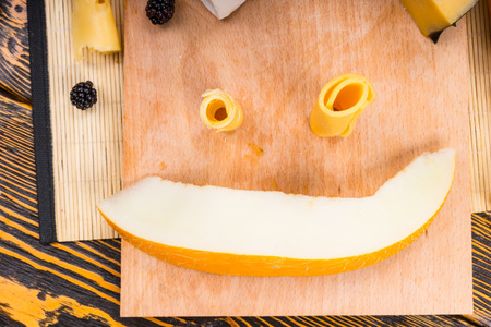 High Angle View of Happy Smiling Face Made from Rolled Cheese Slices and Fruit Wedge on Wooden...