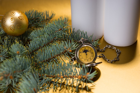 Close Up Festive Still Life of Antique Pocket Watch Surrounded by White Pillar Candles and Evergreen Branch Decorated with Single Gold Christmas Ball on Yellow Backgroundの写真素材