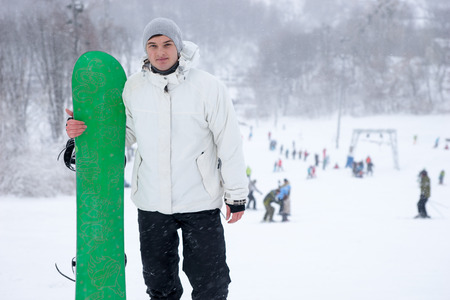 Athletic young man holding a bright green snowboard on a snowy mountain run at a ski resort posing fro the camera with a smileの写真素材