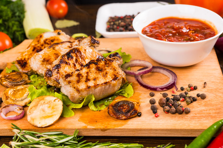 Close Up Still Life View of Grilled Pork or Chicken Chops Resting on Wooden Cutting Board with Bowl of Salsa, Sliced Vegetables and Peppercornsの写真素材