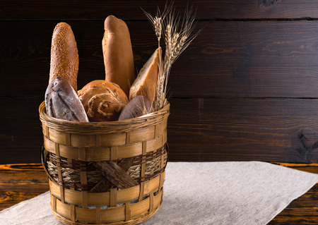 Assorted loaves of bread, rolls and baguettes in a rustic basket on an old wooden table with a wooden background with copy spaceの写真素材