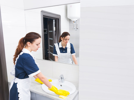 Reflection of female housekeeping worker wearing yellow rubber gloves and white and blue uniform cleaning sink in bathroomの写真素材