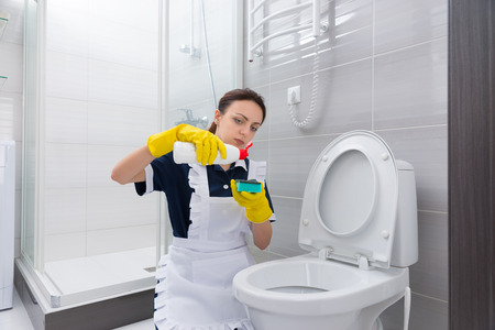 Female housekeeper in blue dress and white apron putting cleaner on green sponge in preparation to scrub down toilet in bathroomの写真素材