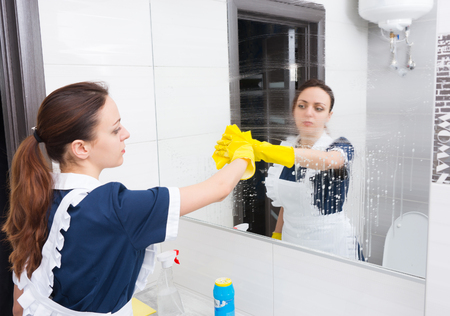 Dirty hotel bathroom mirror being cleaned up with yellow rag by maid in blue and white uniform and...