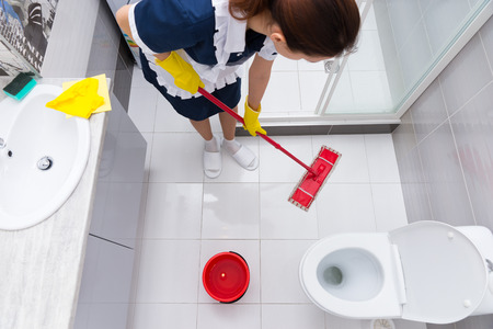 Housekeeper or maid in a neat uniform and apron standing mopping the floor in a bathroom viewed from aboveの写真素材