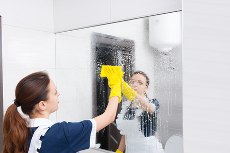 Housekeeper or maid cleaning a large wall mirror in a white bathroom wiping it with a cloth after spraying it with detergentの写真素材
