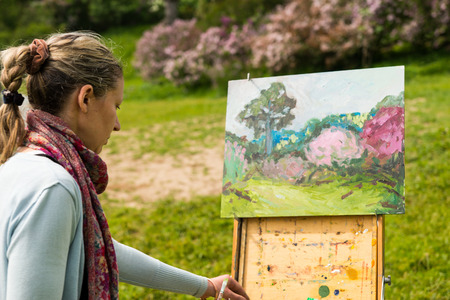 Female painter working outdoors in the park  on a trestle and easel painting with oils and acrylics holding paintbrushの写真素材