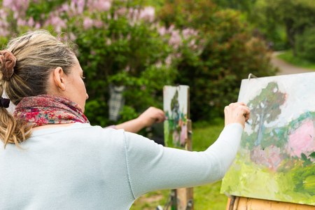 Back view of a woman painter working outdoors in the park or garden on a trestle and easel painting with oils and acrylics during an art classの写真素材