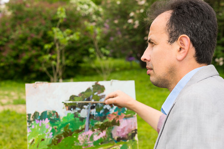 Portrait of a pensive professional painter painting in a forest and holding a paintbrush standing in front of his easelの写真素材