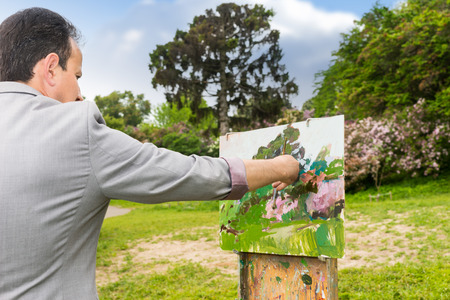 Back view of a man painter working outdoors in the park or garden on a trestle and easel painting with oils and acrylics during an art classの写真素材