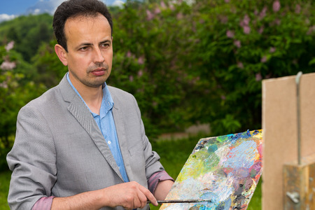 Pensive middle-aged painter holding a palette standing in front of a sketchbook during an art class in a forestの写真素材