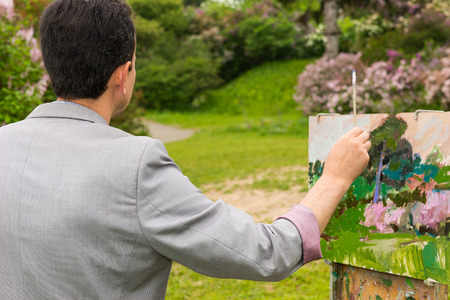 Back view of a male artist with his sketchbook in the park  working  on a trestle and easel painting with oils and acrylics during an art classの写真素材
