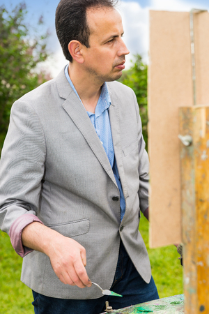 Male handsome middle-aged creative artist working  on a trestle and easel painting with oils and acrylics during an art class in a parkの写真素材
