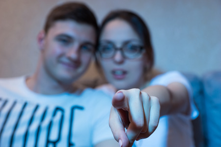 Young girl in glasses is pointing at the camera focus on her finger sitting on a sofa with her boyfriend at home watching television, frontal view in the blue glow from the setの写真素材