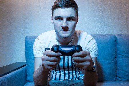 Young man playing a video game with a very serious and concentrated face as he holds his console in...