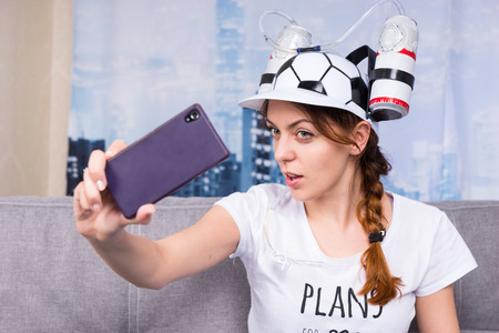 Female football supporter taking selfie by her cell phone in a goofy hat shaped like a football with flasks of liquid on either side sitting on a sofa at homeの写真素材