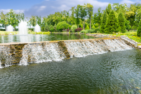 Beautiful waterfall in a park with human made pond, fountain and different trees in a backgroundの写真素材