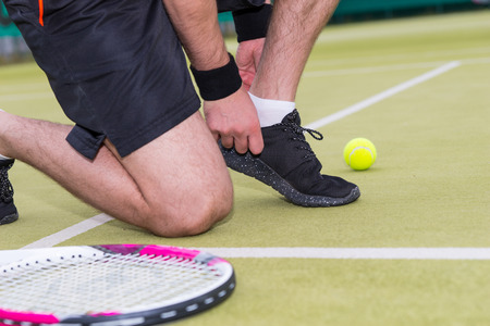Male tennis player tying shoelaces left racket and ball on a court outdoor in summer or spring