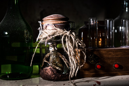 Horrible dead creature with bulging eyes inside jar sealed with string between glass jars and bottles with blood spattered wall in dark room in a Halloween horror conceptの写真素材