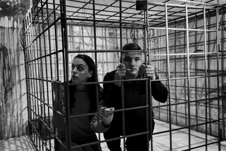 Black and white picture of couple afraid Halloween victims imprisoned in a metal cage with a blood splattered wall behind them looking out through the barsの写真素材