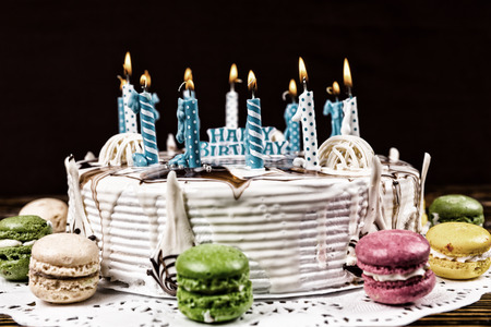 White appetizing birthday cake with lots of burning candles near different colored macarons