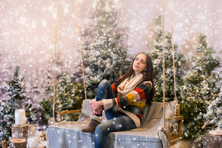 Female dreaming while sitting on a bench or a swing with a blanket under the flashlights in a snow-covered park with spruce trees, wearing warm woolen sweater and knitted scarf while snowingの写真素材