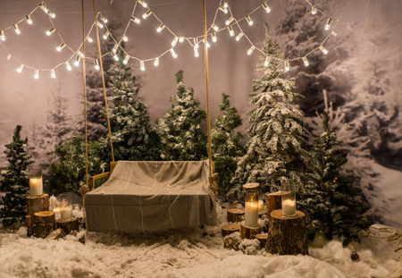 Swing with a blanket on it under the flashlights in a snow-covered park or a forest with spruce trees and stumps with big candles in glass vasesの写真素材