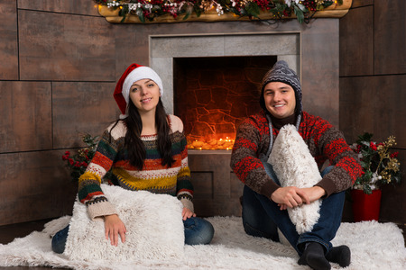 Young smiling couple in warm sweaters and funny winter hats sitting on a rug in front of the decorated fireplaceの写真素材