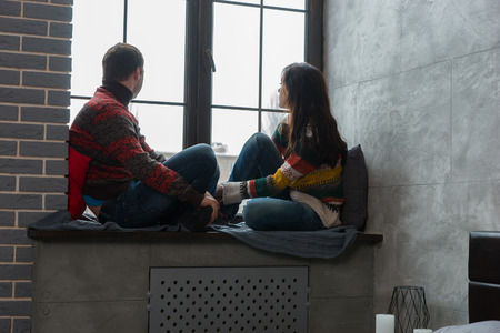 Young couple in warm knitted sweaters looking out the window while sitting on the windowsill with pillows and blanketの写真素材