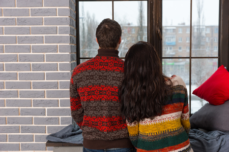 Young pair in warm knitted sweaters looking out the window while standing near windowsill with pillows and blanketの写真素材