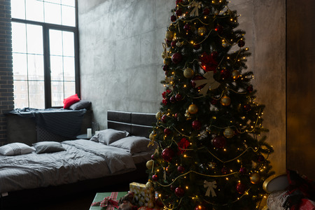 Stylish bedroom with modern bed with a gray bed linen and large Christmas tree with a lot of presents and different decorationsの写真素材