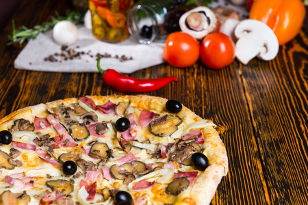 Close up of tasty pizza with ham, onion, olives on wooden table, tomatoes, mushrooms and other vegetables in the backgroundの写真素材