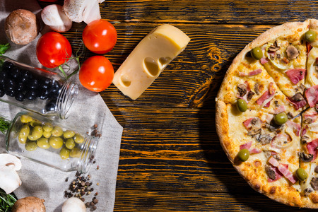 High angle view of half of pizza with ham, onion, olives, near lie tomatoes, cheese, mushrooms and other vegetables on napkin on wooden tableの写真素材