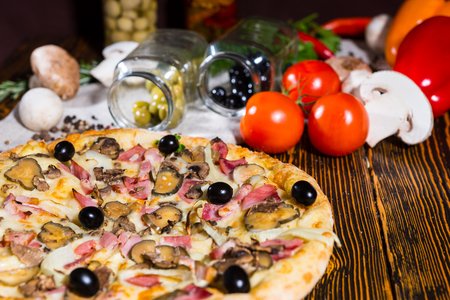Tasty pizza with black olives on wooden table, tomatoes, mushrooms and other vegetables in the backgroundの写真素材