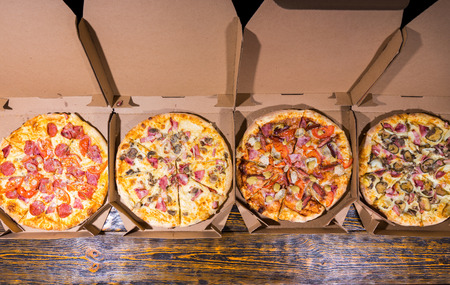 High angle view of four pizzas with variety of toppings and cheese in cardboard take out boxes with open lid on wooden tableの写真素材
